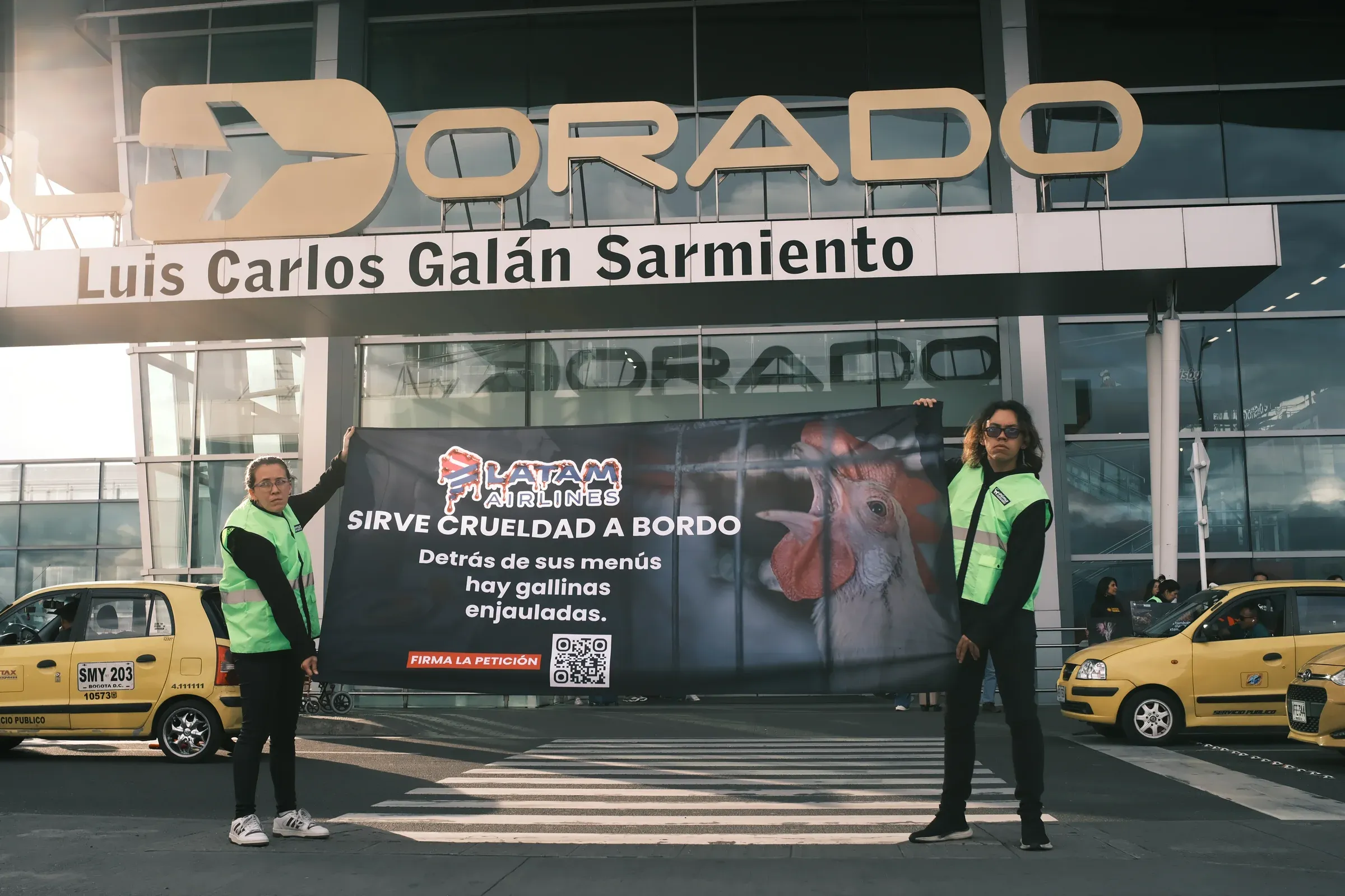 Dos activistas con chaleco sostienen frente a la entrada del Aeropuerto El Dorado una pancarta que dice «LATAM Airlines sirve crueldad a bordo. Detrás de sus menús hay gallinas enjauladas».